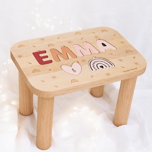 Könnte beinhalten: Ein heller Holzhocker mit vier zylindrischen Beinen. Die Oberseite zeigt den Namen EMMA in ausgeschnittenen Buchstaben mit Farbverläufen in Rot, Orange und Rosa sowie ein Herz und ein Regenbogenmotiv mit kleinen eingravierten Symbolen.