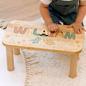 May include: A light-coloured wooden step stool featuring a custom name puzzle with the letters W, I, L, A, I, A, M in various pastel colours. The surface is decorated with engraved dinosaur and palm tree illustrations, providing a functional surface.