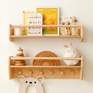 Op de afbeelding: Twee lichtgekleurde houten wandplanken met veiligheidsrails aan de voorzijde. De bovenste plank bevat drie boeken en een glazen pot. De onderste plank bevat een houten stapelspeelgoed, een houten regenboog, een pluche konijn en zes stervormige haken.