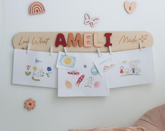 以下が含まれることがあります： 「Look What AMELI Made」の文字が赤と茶色で書かれた木製のディスプレイボード。子供たちの絵を小さなクリップで留めています。虹、蝶、ハート、花の装飾的な木製シェイプが壁に取り付けられています。