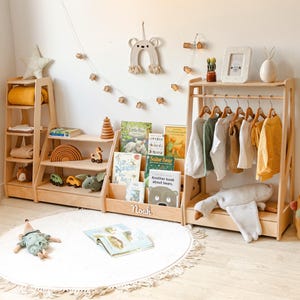 May include: A light-colored wooden modular storage unit featuring a tiered bookshelf, a clothing rack with hangers, and open shelving. The unit displays various children's books, toys, and hanging garments, with the name Noah printed on the front.