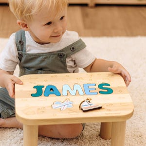 May include: A light-colored wooden stool featuring a personalized name puzzle with the letters J-A-M-E-S in shades of blue. The surface is decorated with engraved cloud and wave patterns, and includes two small wooden puzzle pieces shaped like a plane and a boat.