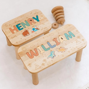 May include: Two light-coloured wooden stools with four legs each. The smaller stool features the name HENRY in colourful letters with a bear and mountain graphic. The larger stool features the name WILLIAM with dinosaur graphics. Both have engraved nature motifs.