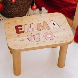 May include: A light-colored wooden stool featuring the name EMMA in cutout letters with varying shades of pink and red. The surface is decorated with engraved butterfly and flower motifs, including a pink butterfly and a pink flower cutout.