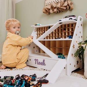 Può includere: Un organizer per auto giocattolo in legno bianco con più scomparti e una rampa. L'organizer ha ritagli a forma di stella e il nome "Noah" in scrittura viola. Una collezione di auto giocattolo è sparsa sul pavimento.