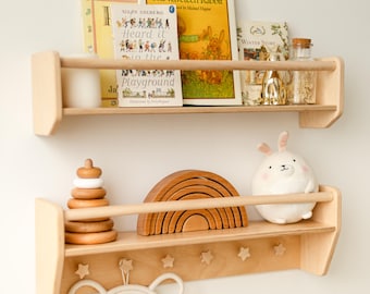 May include: Two light-colored wooden wall shelves with front safety rails. The top shelf holds books titled The Velveteen Rabbit and Heard it in the Playground. The bottom shelf features a wooden stacking toy, a rainbow arch, and a plush rabbit toy.