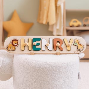 May include: Wooden name puzzle spelling "HENRY" with colorful letters and animal illustrations. The puzzle features a lion and giraffe, with a light wood finish and wooden pegs for each letter. The name puzzle is a children's toy.