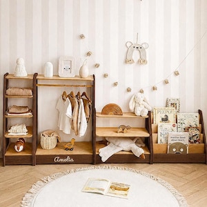 May include: A dark wood modular children's furniture set featuring a four-tier shelving unit, a hanging garment rack with the name Amelia engraved on the base, a two-tier storage shelf, and a book display rack, all arranged against a striped wall.