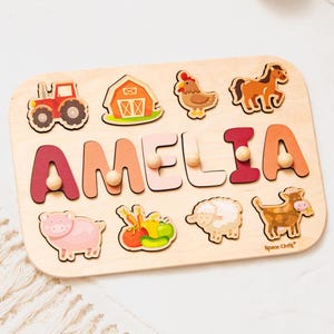May include: Wooden puzzle featuring the name "AMELIA" in colorful letters with wooden pegs. The puzzle includes farm animal shapes like a tractor, barn, chicken, horse, pig, and cow. The puzzle is made of light-colored wood.