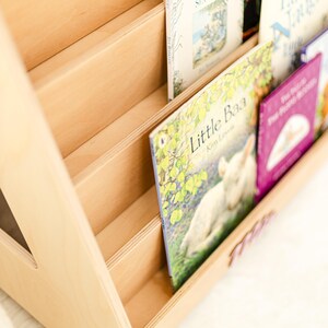 May include: A light-colored wooden bookshelf with multiple slanted tiers for displaying books. The shelves hold several children's books, including one titled Little Baa with an illustration of a lamb, and a purple book with white text on the cover.