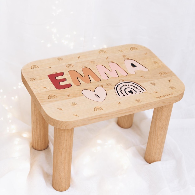 Puede incluir: Un taburete de madera clara con cuatro patas cil&iacute;ndricas y una superficie rectangular. La parte superior muestra el nombre EMMA en letras recortadas con degradado de color, un coraz&oacute;n y un arco&iacute;ris, junto a peque&ntilde;os iconos grabados.