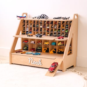 May include: A light-colored wooden toy storage shelf with multiple grid compartments for small vehicles. The unit features a ramp on the right side and the name Noah in white script lettering on the front panel. It is filled with various toy cars.