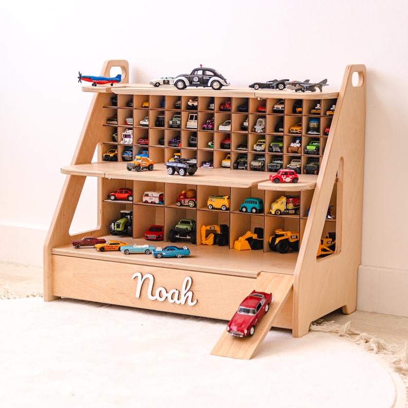 May include: A light-colored wooden toy storage shelf with multiple grid compartments for small vehicles. The unit features a ramp on the right side and the name Noah in white script lettering on the front panel. It is filled with various toy cars.