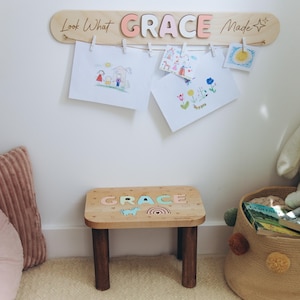 Może przedstawiać: Drewniana tablica z napisem "Look What GRACE Made" i pasujący drewniany stołek. Tablica zawiera grafiki z małymi spinaczami. Stołek ma imię "GRACE" oraz wz&oacute;r tęczy i jednorożca.