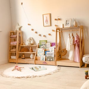 May include: A light-colored wooden nursery furniture set including a tall shelving unit, a tiered bookshelf with the name Ellie, and a clothing rack with hangers. The setup features various children's books, a woven bag, and hanging garments.