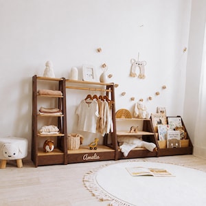 May include: A dark wood modular storage unit featuring a tall shelving section, a central clothing rack with a hanging rod, and a low tiered bookshelf. The central base is personalized with the name Amelia in white script lettering.