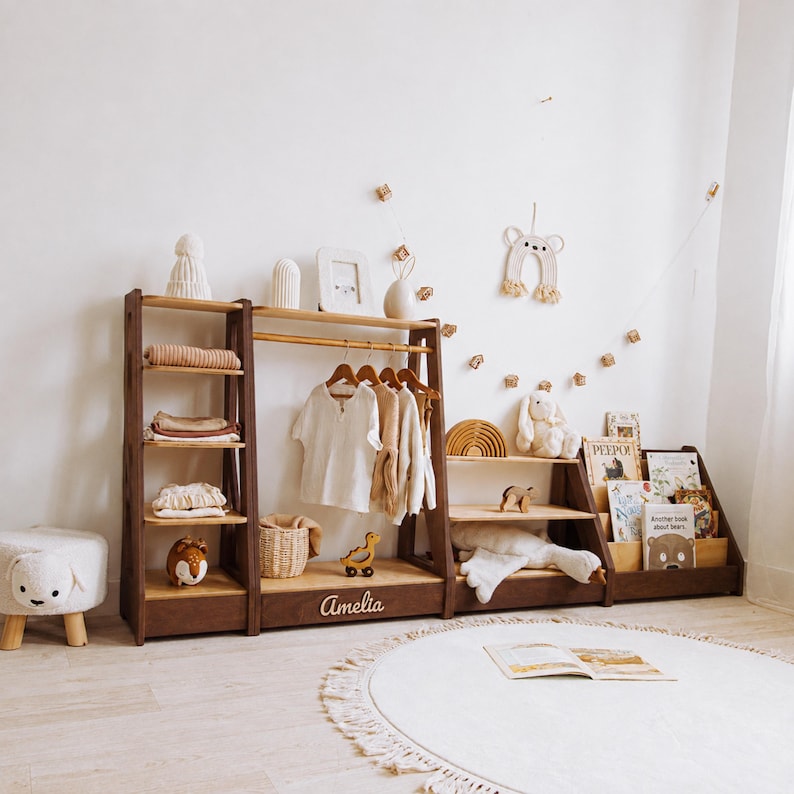 May include: A dark wood modular storage unit featuring a tall shelving section, a central clothing rack with a hanging rod, and a low tiered bookshelf. The central base is personalized with the name Amelia in white script lettering.