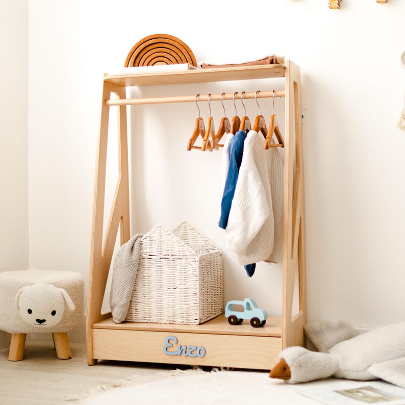 May include: A light-colored wooden clothing rack with a top shelf, a hanging rod with six wooden hangers, and a bottom drawer featuring the name Enzo in blue script. A white woven basket sits on the bottom shelf next to a small blue toy truck.
