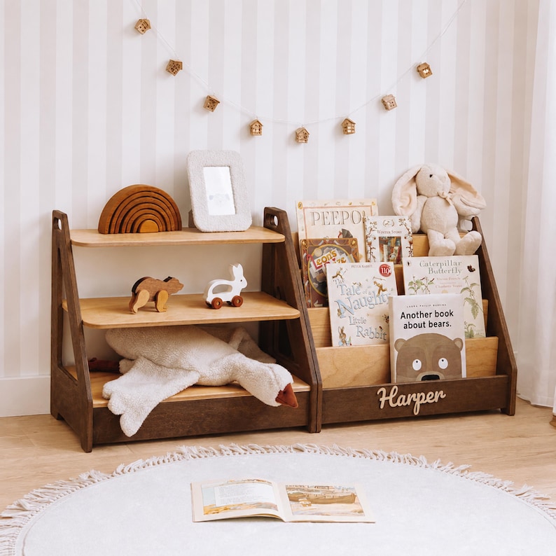 May include: A wooden toy storage unit with three shelves and a book display section. The unit features a dark brown frame with light wood shelves. The name Harper is displayed in white script on the front. It holds books, a plush rabbit, and toys.