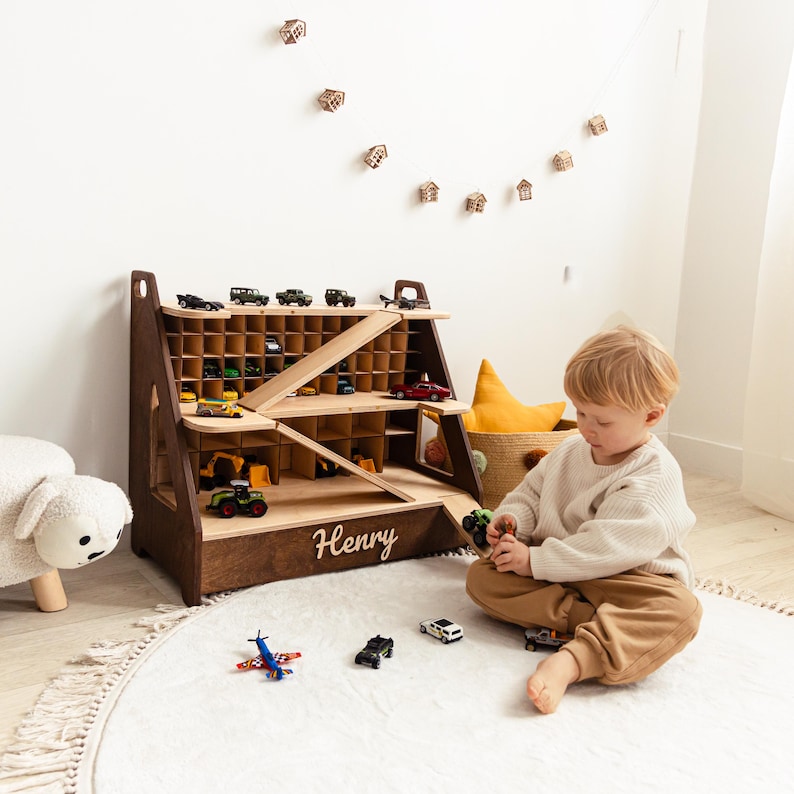 May include: A dark brown wooden toy car storage garage with multiple cubby compartments, two diagonal ramps, and the name Henry in white script lettering on the front base. The structure holds various small toy vehicles on its shelves.