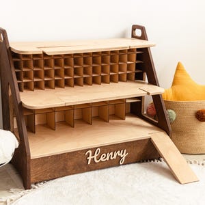 May include: A two-tiered wooden display shelf with a dark brown frame and light wood shelves. The top tier features a grid of small square compartments, while the bottom tier has larger dividers. The name Henry is attached to the front base.