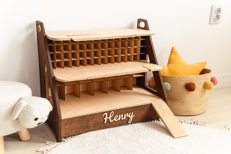 May include: A two-tiered wooden display shelf with a dark brown frame and light wood shelves. The top tier features a grid of small square compartments, while the bottom tier has larger dividers. The name Henry is attached to the front base.