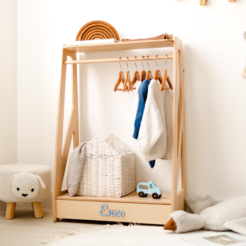May include: A light-colored wooden clothing rack with a top shelf, a hanging rod with six wooden hangers, and a bottom drawer featuring the name Enzo in blue script. A white woven basket sits on the bottom shelf next to a small blue toy truck.