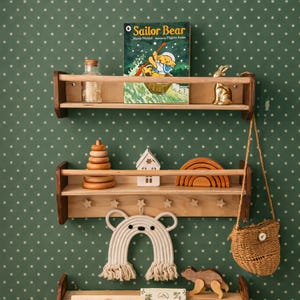 May include: Two light-colored wooden wall shelves with front rails. The top shelf holds a Sailor Bear book, a glass jar, and a gold rabbit figurine. The bottom shelf features a wooden stacking toy, a house, a rainbow, and star-shaped hooks.