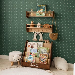 May include: A dark wood floor book rack with the name Amelia engraved on the front, holding several children's books. Above it are two wall-mounted wooden shelves displaying books, a wooden stacking toy, a house, a rainbow, and a woven bag.