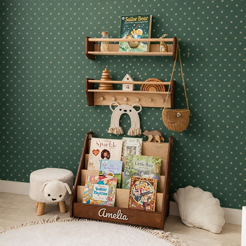 May include: A dark wood floor book rack with the name Amelia engraved on the front, holding several children's books. Above it are two wall-mounted wooden shelves displaying books, a wooden stacking toy, a house, a rainbow, and a woven bag.