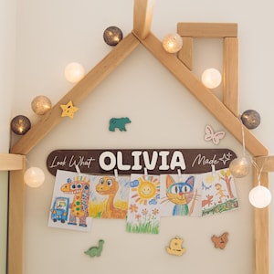 Może przedstawiać: Drewniana rama w kształcie domu z łańcuchem świetlnym i grafikami dla dzieci. Brązowy znak głosi "Look What OLIVIA Made". Wyświetlane są kolorowe rysunki zwierząt i słońca. Ozdobne drewniane kształty: gwiazda, niedźwiedź, motyl, dinozaur, ł&oacute;dź i gwiazda.