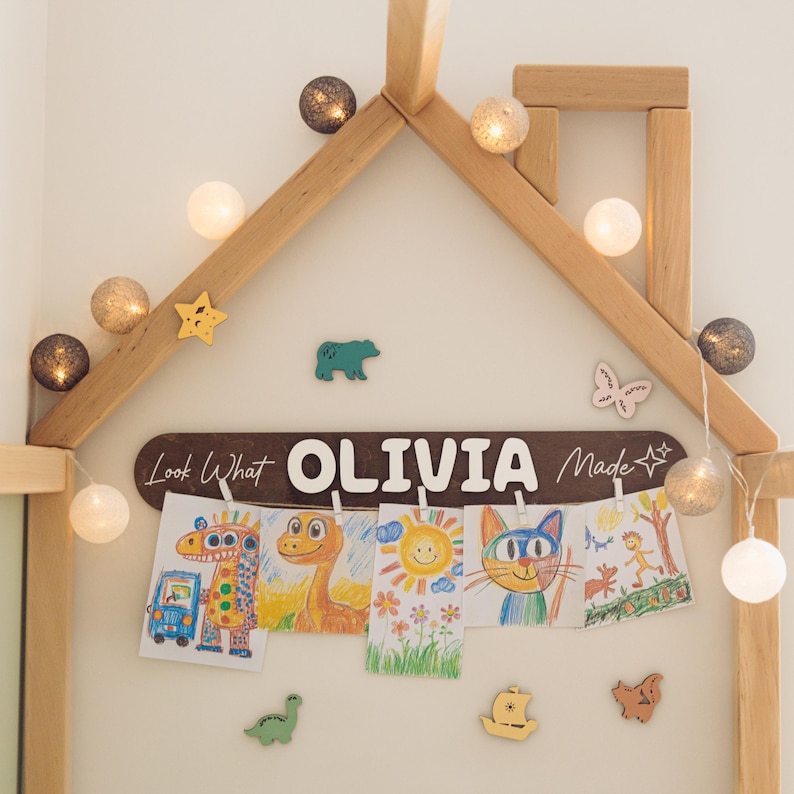 Może przedstawiać: Drewniana rama w kształcie domu z łańcuchem świetlnym i grafikami dla dzieci. Brązowy znak głosi "Look What OLIVIA Made". Wyświetlane są kolorowe rysunki zwierząt i słońca. Ozdobne drewniane kształty: gwiazda, niedźwiedź, motyl, dinozaur, ł&oacute;dź i gwiazda.