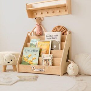 May include: A light-colored wooden bookshelf with four tiered shelves, featuring the name Naomi in white script on the front. The shelf holds several children's books, including titles like Seymour and Henry and Another book about bears.
