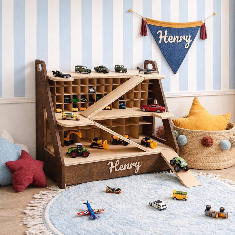 May include: A dark wood toy storage shelf with a grid of cubbies, two diagonal ramps, and the name Henry in white script on the front. The shelf holds various miniature toy cars, trucks, and construction vehicles, with more scattered on the floor.