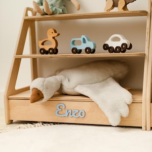 May include: A light-colored wooden bookshelf with three tiers. The middle shelf holds a wooden duck, a light blue truck, and a white car. The bottom shelf features a plush white goose toy and the name Enzo in light blue script lettering.