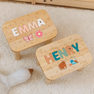 May include: Two rectangular wooden step stools with rounded corners. One stool features the name EMMA in white letters with a pink butterfly and flower. The other stool features the name HENRY in teal and orange letters with a brown bear and mountain graphic.
