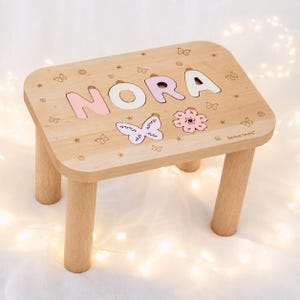 Puede incluir: Un taburete de madera clara con cuatro patas cil&iacute;ndricas y una superficie rectangular. La parte superior muestra el nombre NORA en letras de color rosa pastel y blanco, junto con motivos grabados de mariposas y flores y el texto Space Girls.