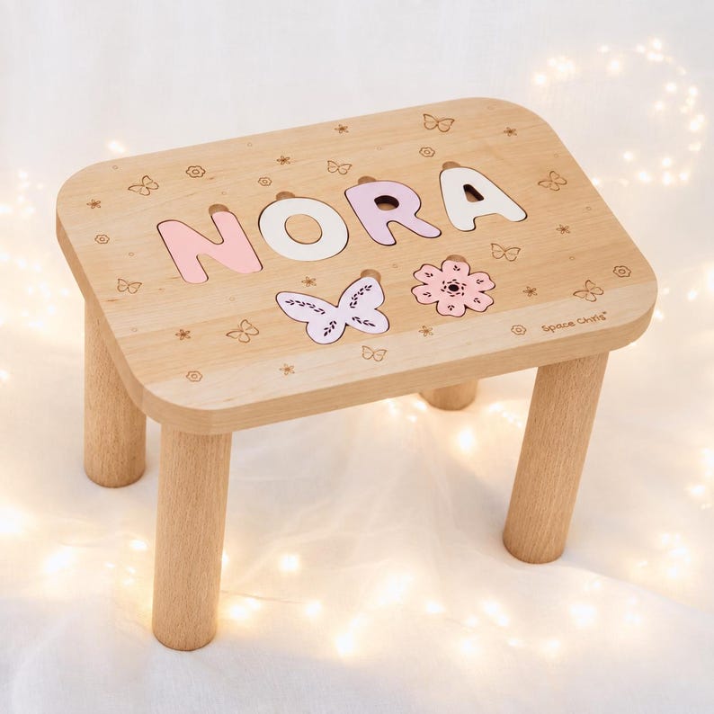 Puede incluir: Un taburete de madera clara con cuatro patas cil&iacute;ndricas y una superficie rectangular. La parte superior muestra el nombre NORA en letras de color rosa pastel y blanco, junto con motivos grabados de mariposas y flores y el texto Space Girls.