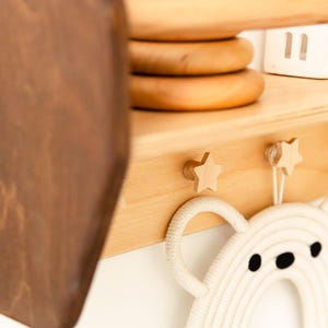 May include: A light-colored wooden wall shelf featuring two star-shaped knobs and a dark brown side panel. A white rope-woven bear-shaped decoration hangs from the star knobs, while stacked wooden rings sit on the top shelf surface.