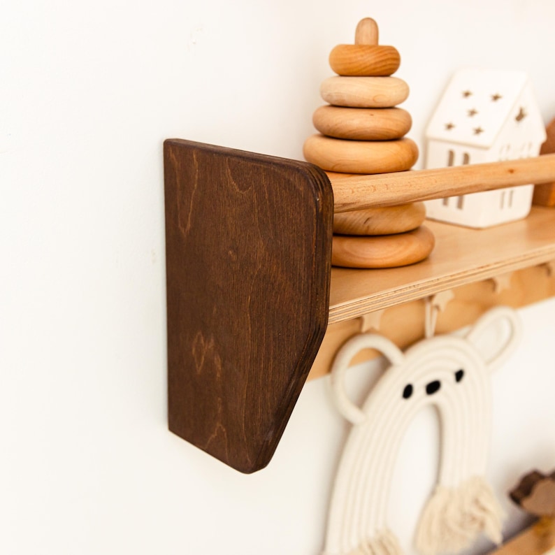 May include: A wall-mounted wooden shelf with a dark brown side panel and a natural light wood finish. The shelf features a horizontal front rail and is decorated with a wooden stacking ring toy, a white house-shaped ornament, and a macrame bear.