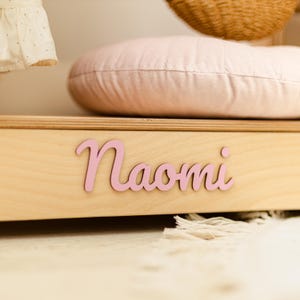May include: A light-colored wooden shelf featuring the name Naomi in pink, cursive, raised lettering attached to the front edge. A soft, pale pink quilted cushion sits on top of the wooden surface, partially obscuring the view behind it.