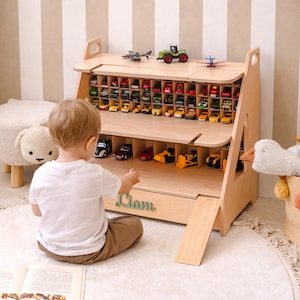 May include: A light-colored wooden toy storage shelf featuring multiple small compartments for miniature cars, a larger lower level for bigger vehicles, a fold-down ramp, and the name Liam in green script lettering on the front panel.