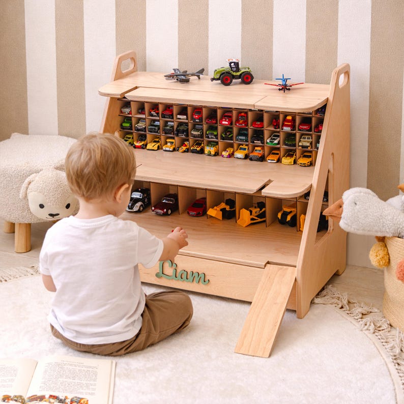 May include: A light-colored wooden toy storage shelf featuring multiple small compartments for miniature cars, a larger lower level for bigger vehicles, a fold-down ramp, and the name Liam in green script lettering on the front panel.