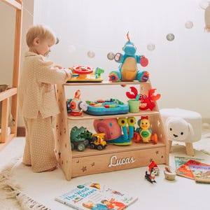 May include: A wooden toy shelf with three tiers, filled with colorful toys, including a blue dinosaur, a red crab, and a yellow duck. The shelf has the name "Lucas" on the front. A child in pajamas stands near the shelf.