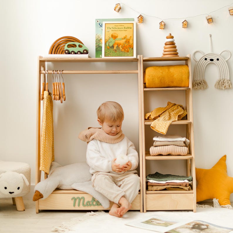 May include: A light-colored wooden clothing rack with a hanging rod and attached shelving unit. The rack features a base with the name Mateo in green script. The shelves hold folded yellow and neutral textiles, a wooden stacking toy, and books.