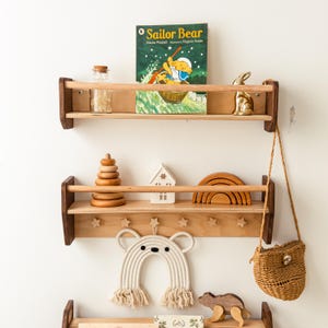 May include: Two wooden wall-mounted shelves with front safety rails. The top shelf holds a Sailor Bear book, a glass jar, and a gold rabbit figurine. The bottom shelf features star-shaped hooks, a wooden stacking toy, a house, and a rainbow arch.