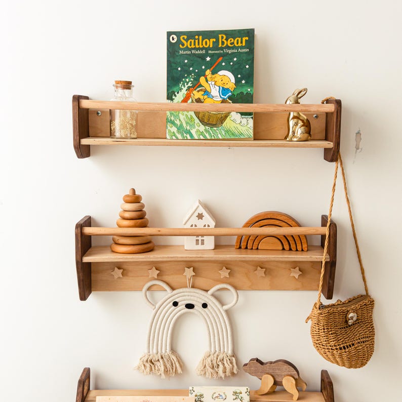 May include: Two wooden wall-mounted shelves with front safety rails. The top shelf holds a Sailor Bear book, a glass jar, and a gold rabbit figurine. The bottom shelf features star-shaped hooks, a wooden stacking toy, a house, and a rainbow arch.