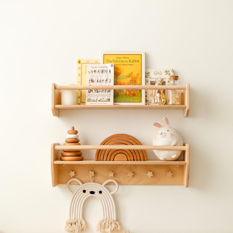 May include: Two light-colored wooden wall shelves with front safety rails. The top shelf holds three books and a glass jar. The bottom shelf features a wooden stacking toy, a wooden rainbow arch, a plush rabbit, and six star-shaped hooks.