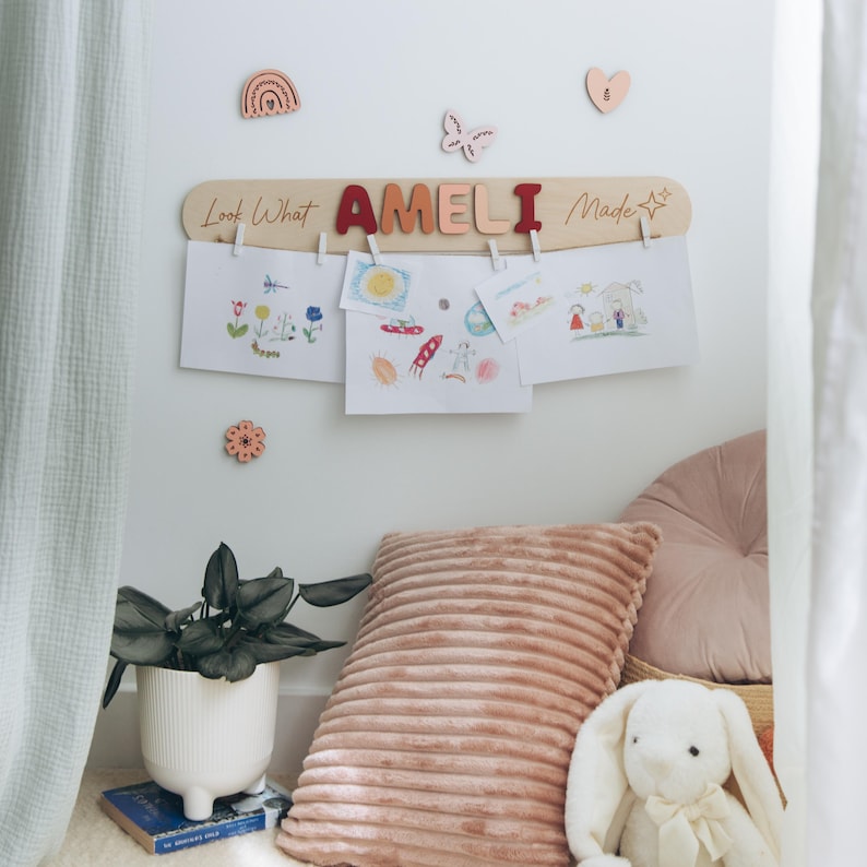Może przedstawiać: Drewniana tablica z napisem "Look What AMELI Made" w czerwonych i pomarańczowych literach, trzymająca rysunki dziecięce za pomocą spinaczy. Ozdobne drewniane kształty, w tym tęcza, motyl, kwiat i serce, są zamontowane na ścianie. Biała donica z rośliną i r&oacute;żowa poduszka znajdują się na pierwszym planie.