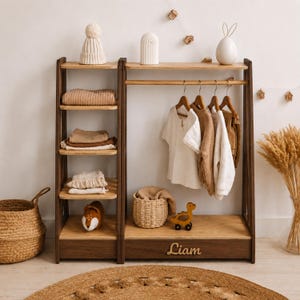 May include: A wooden clothing rack and shelving unit featuring a dark brown frame with light wood shelves. The unit includes a hanging rod with wooden hangers, a bottom drawer with the name Liam in script, and various folded garments and toys.